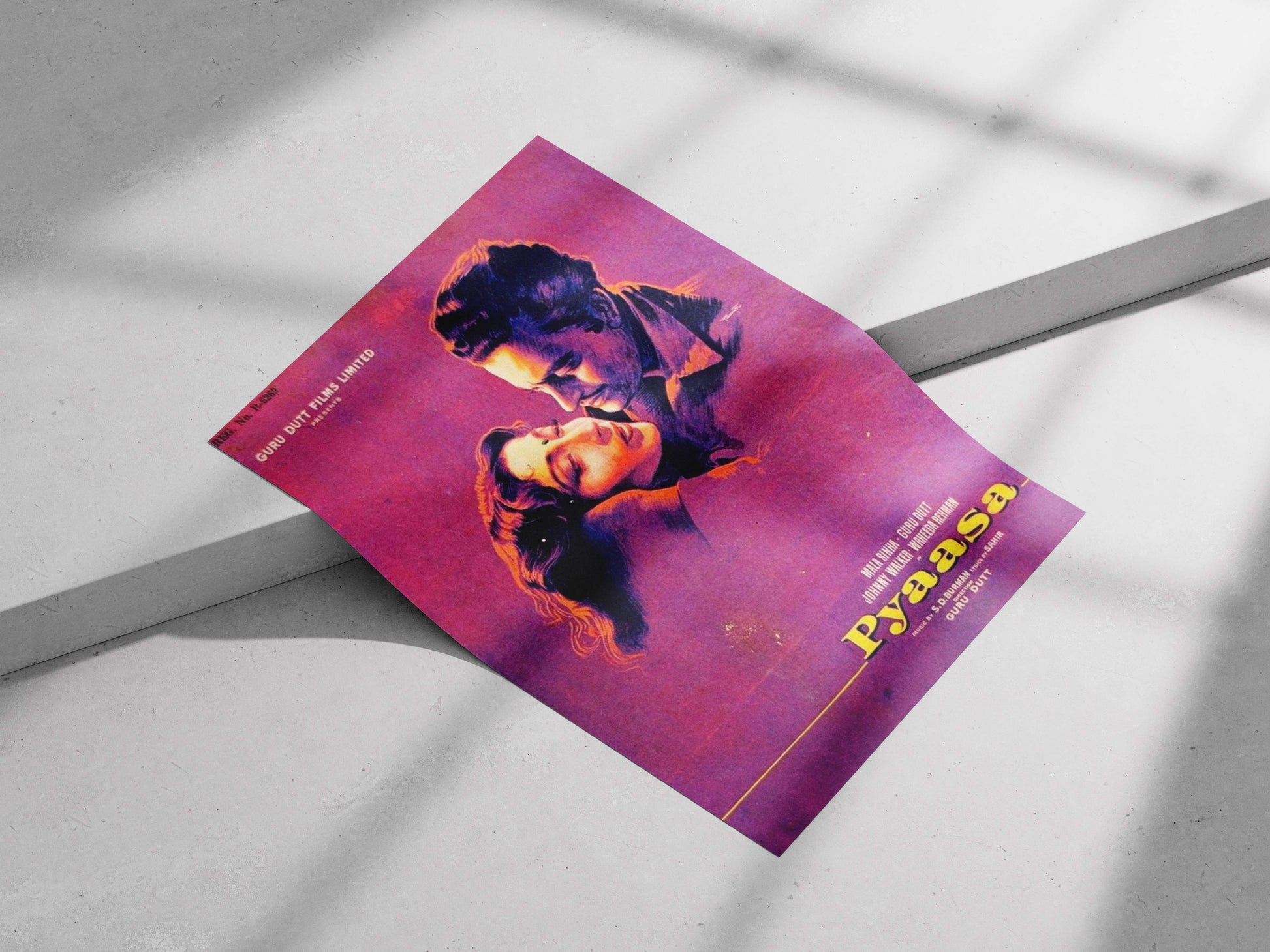 Purple movie poster with a stylized face design on a light gray surface. Retro Pyaasa movie poster featuring Guru Dutt and Waheeda Rehman in a moody purple and orange color scheme, displayed on a studio wall.